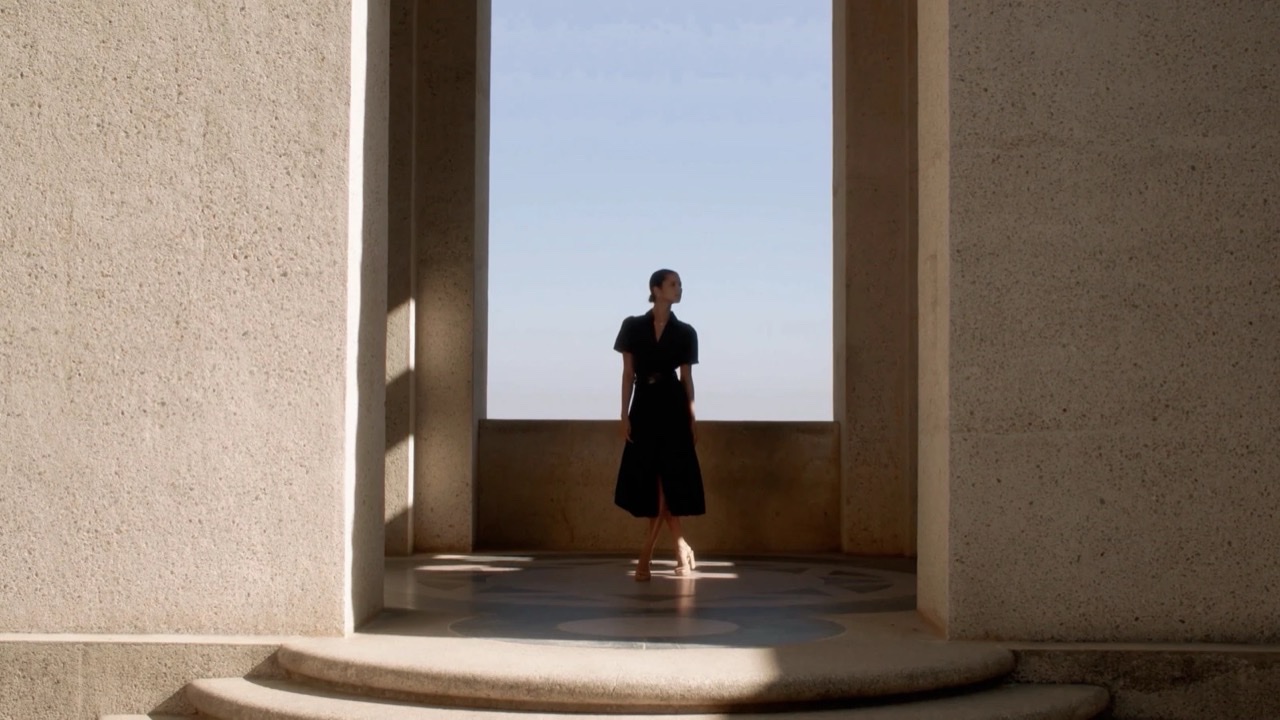 A woman in a black dress walking a sunlit stone colonnade above the sea
