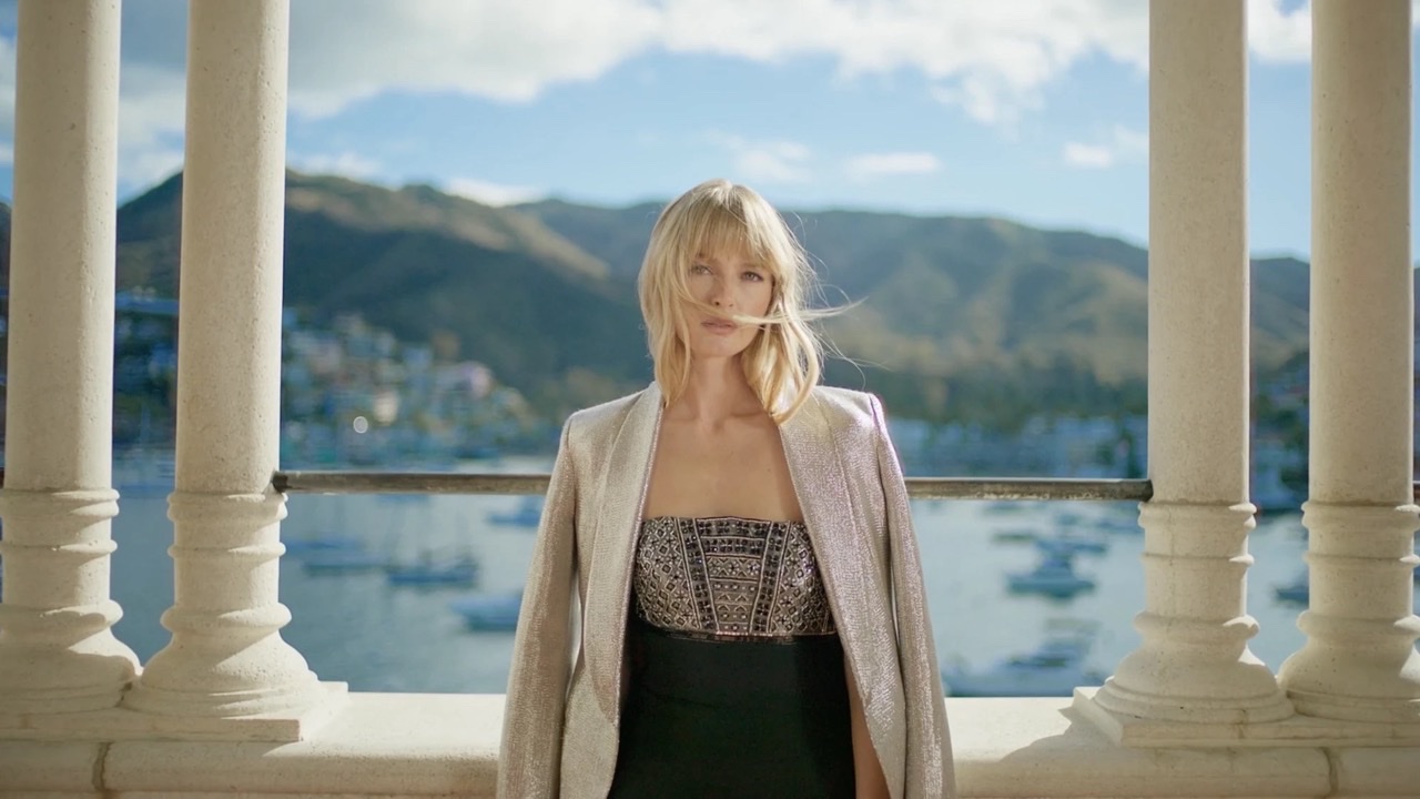 A woman in a beaded gown and shimmer jacket on a colonnaded terrace above a harbour