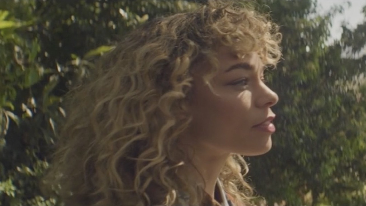 A woman with curly blonde hair turning in sunlight, trees behind her