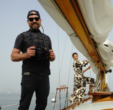 Jeffrey with a camera on the deck of a sailboat, a model at the mast