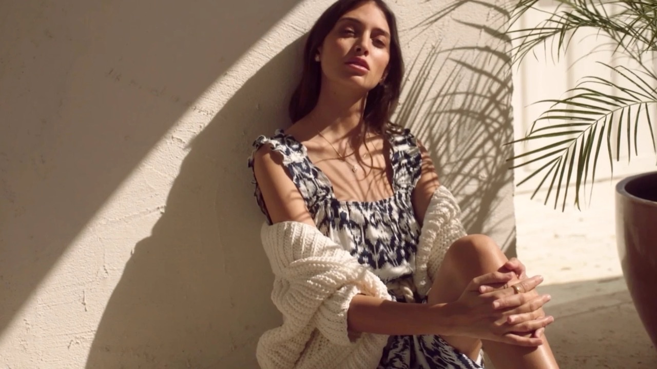 A woman in a printed dress and cream knit cardigan against a sunlit wall, palm shadow across it
