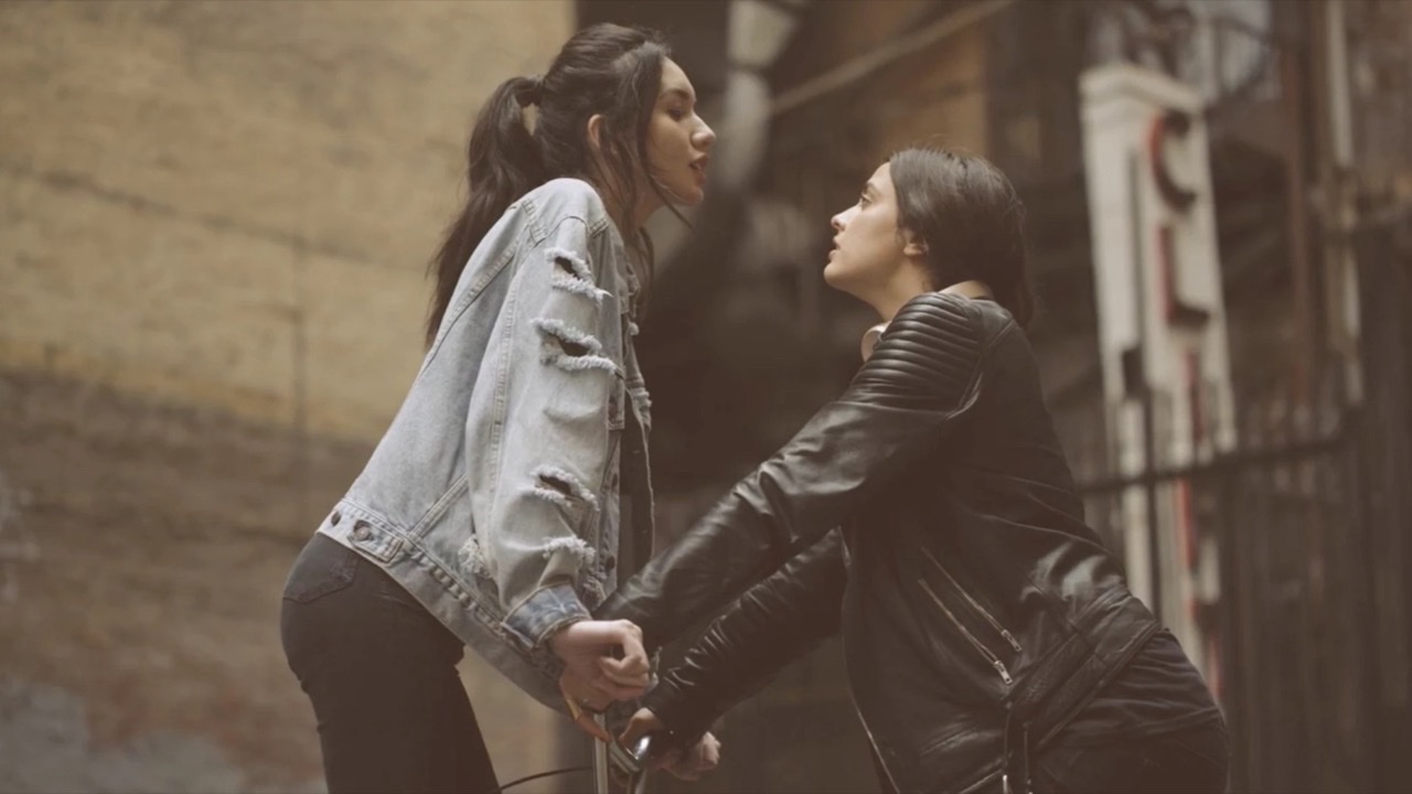 Two women face to face over a bicycle in an alley, denim and leather